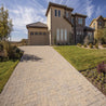 Tan paver stones create a driveway leading to a beige house with green lawn.
