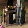 Outdoor kitchen with built-in grill, stone base, and man grilling.
