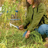 Person kneeling in garden using a billhook saw to trim weeds.
