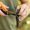 Close-up of hands using sharp garden shears with an orange handle, being stored in a black sheath.
