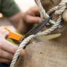 Person using sharp garden shears to cut thick rope near burlap sack.
