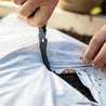 Hands using black gardening tool to cut a hole in white landscape fabric above dark soil.
